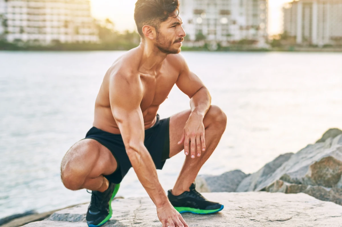 A man squats on a rocky surface near water, wearing shorts and athletic shoes