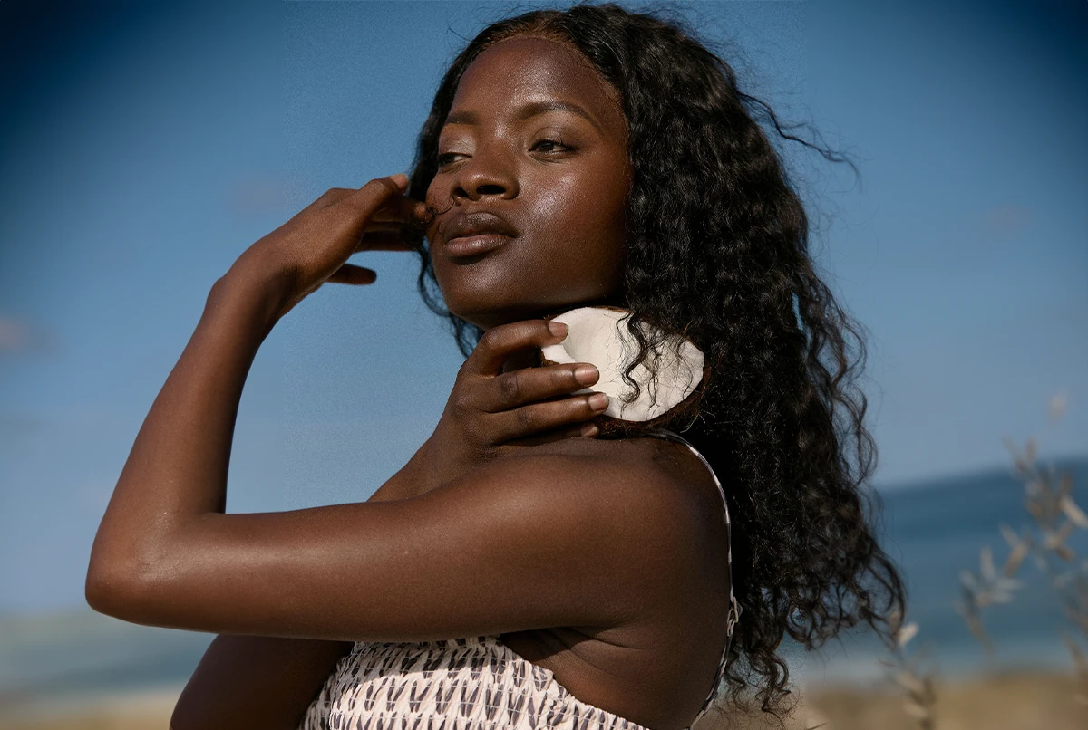 A woman with long, curly hair poses outdoors, holding a shell near her neck