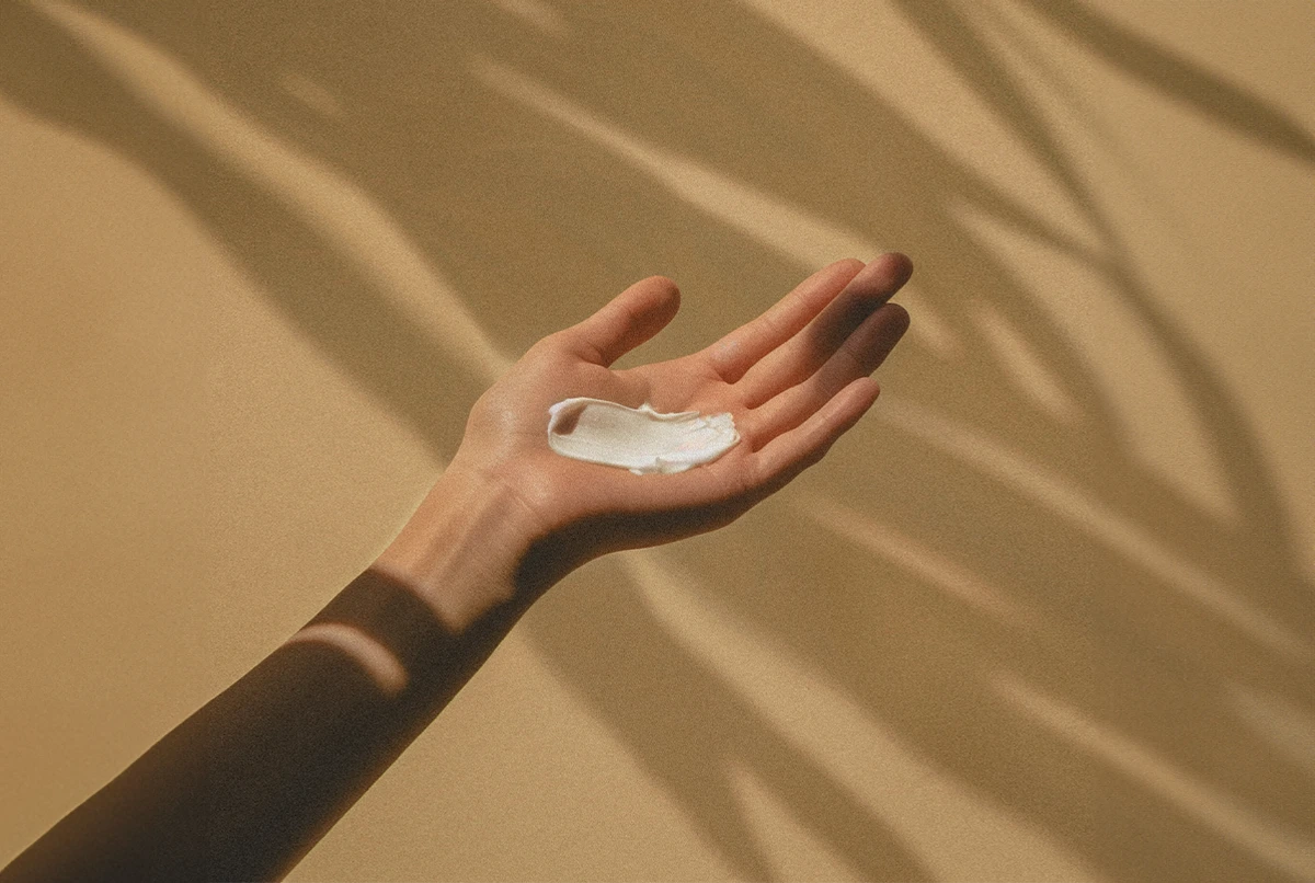 A hand holds a small amount of cream, with soft shadows in the background