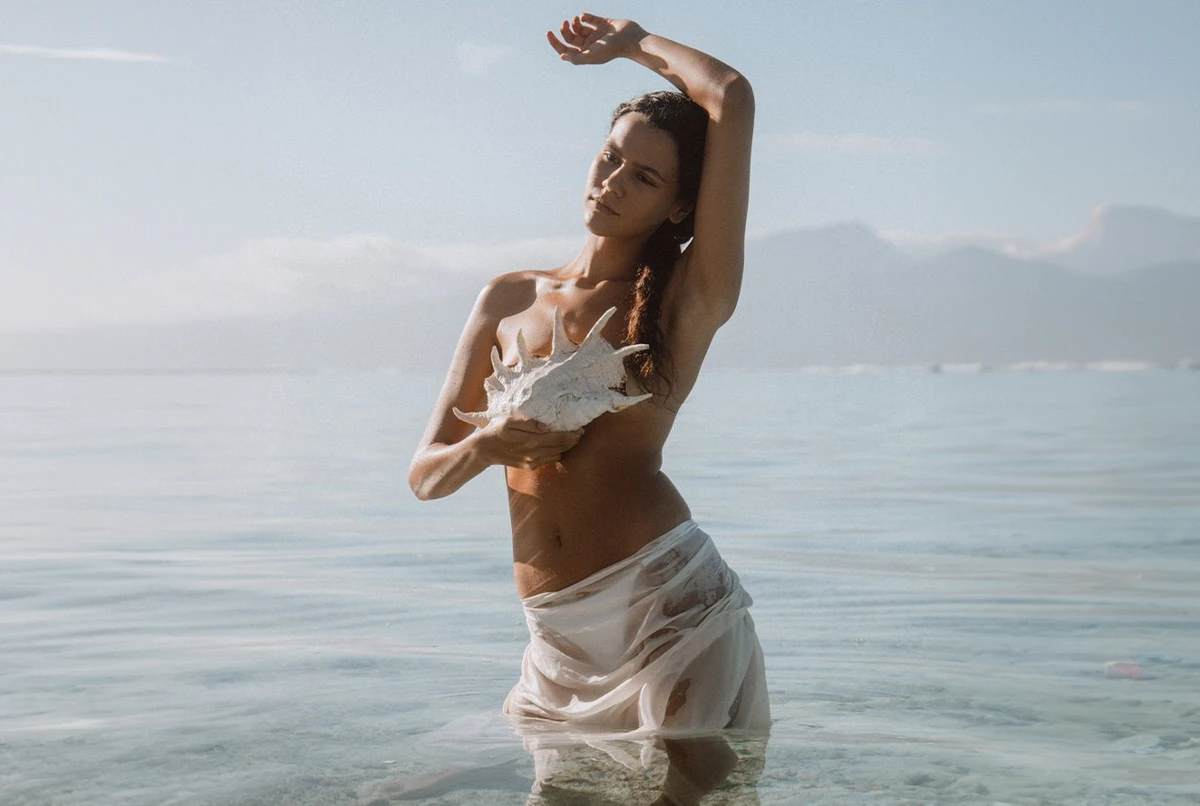 A woman stands in shallow water holding a large shell, wearing a sheer white wrap