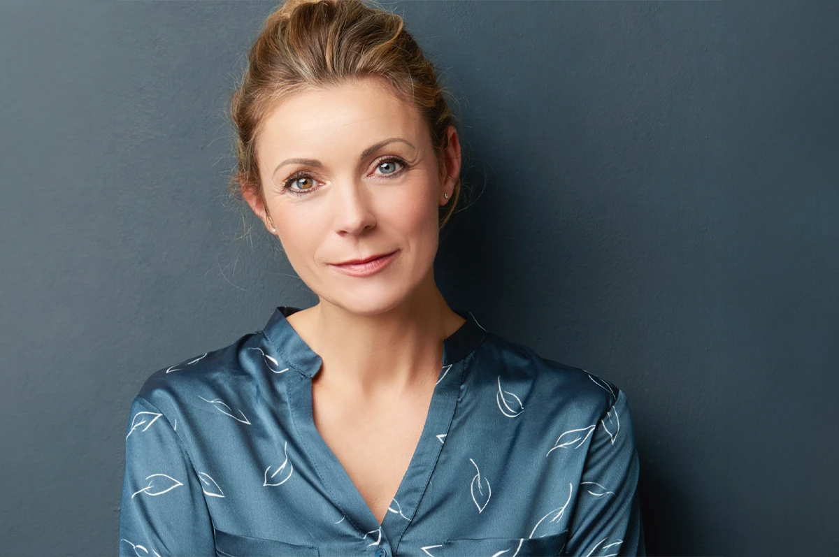 Woman with light brown hair, wearing a blue patterned blouse, standing against a dark wall