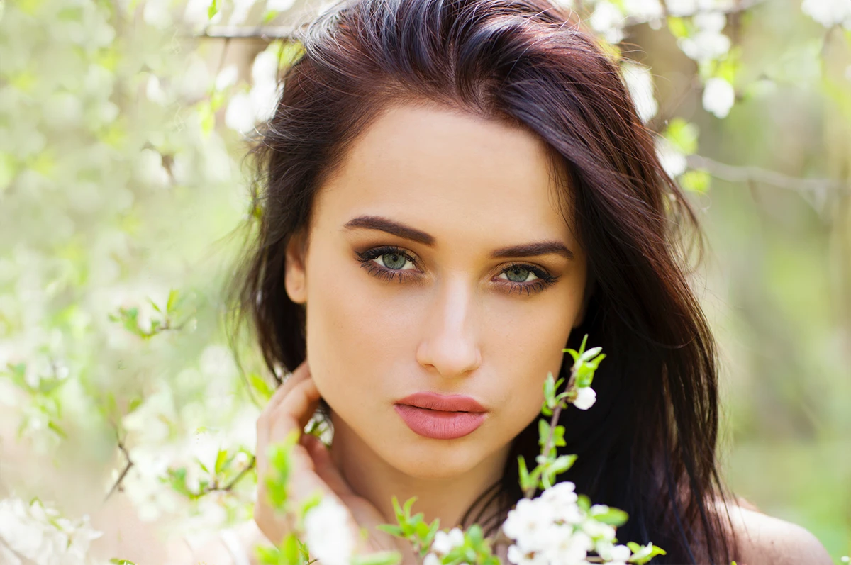 A woman with long dark hair stands amidst flowering branches, gazing at the camera