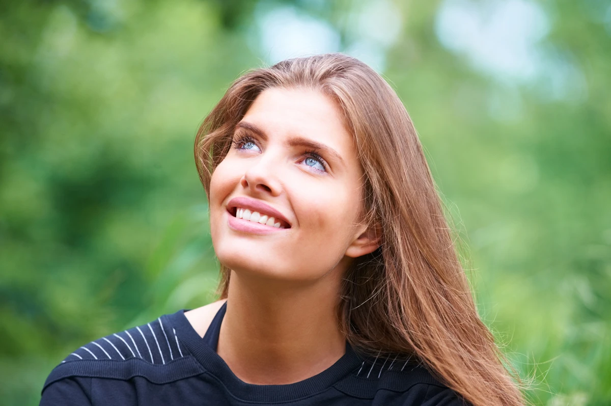A young woman with long brown hair looks up, smiling, in a green outdoor setting