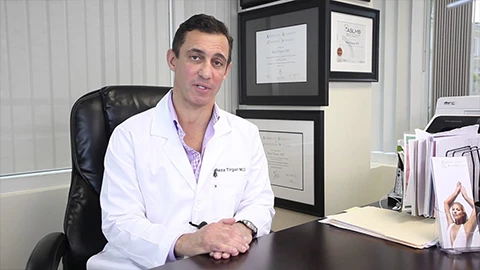A man in a lab coat sits at a desk with certificates on the wall behind him