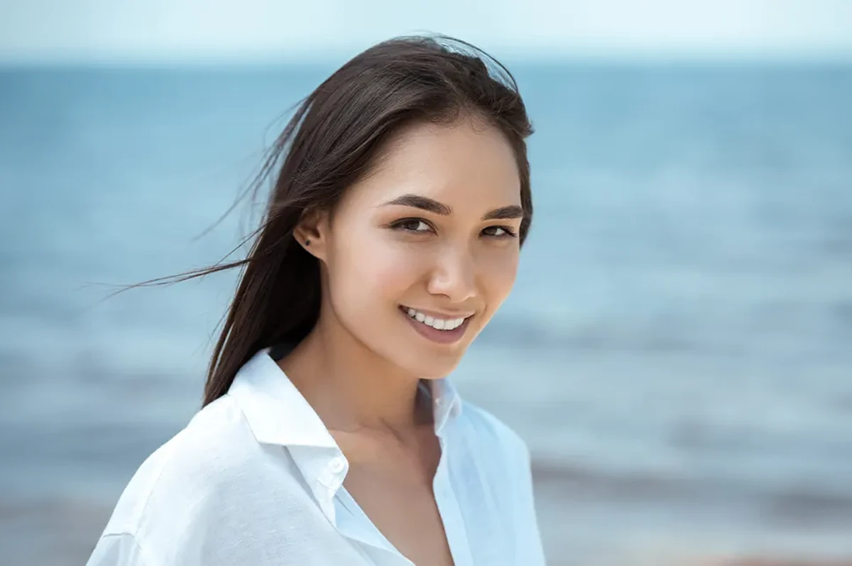 A woman with long dark hair smiles at the beach, with sea in the background