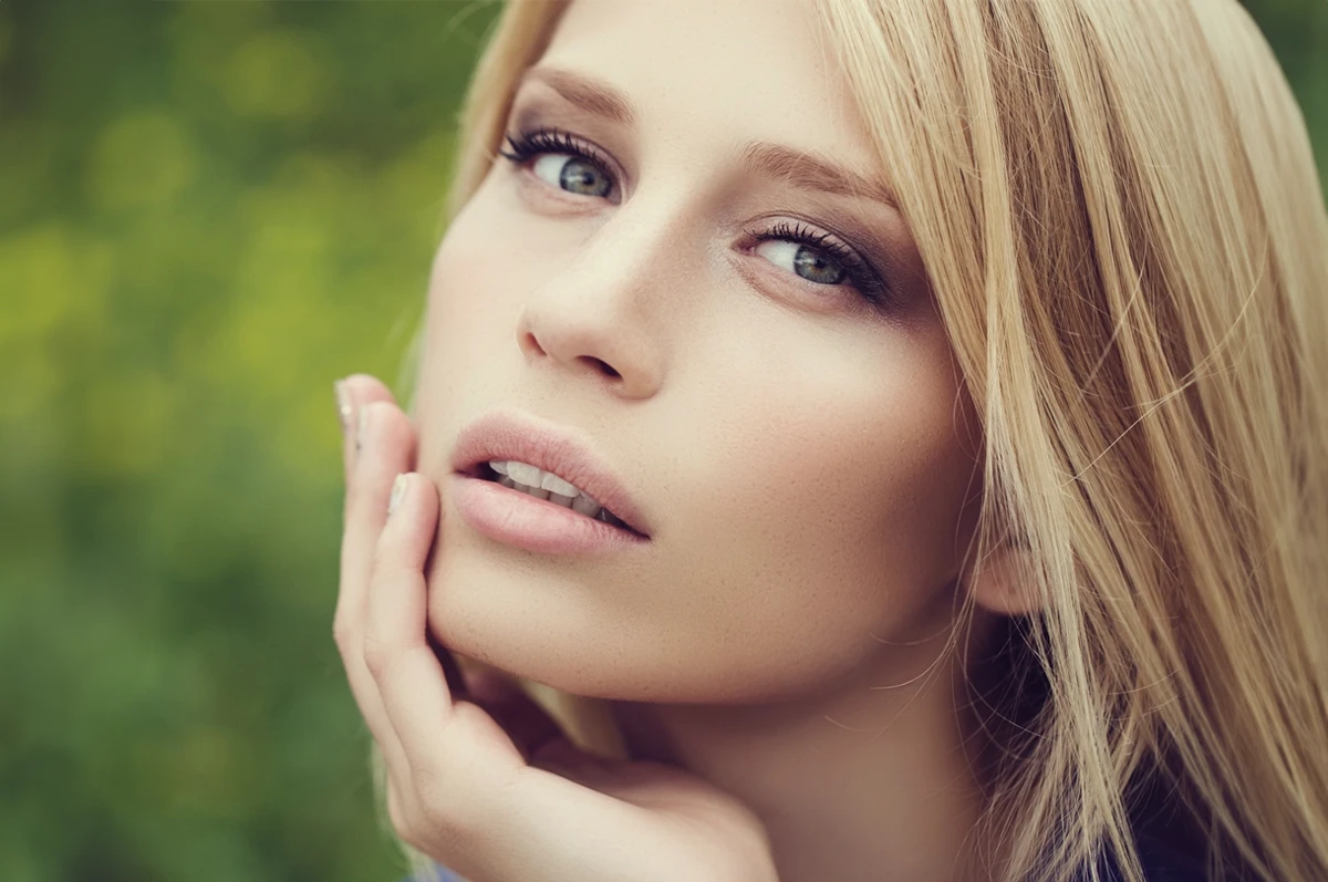 Close-up of a woman with long blonde hair, resting her chin on her hand, against a green background