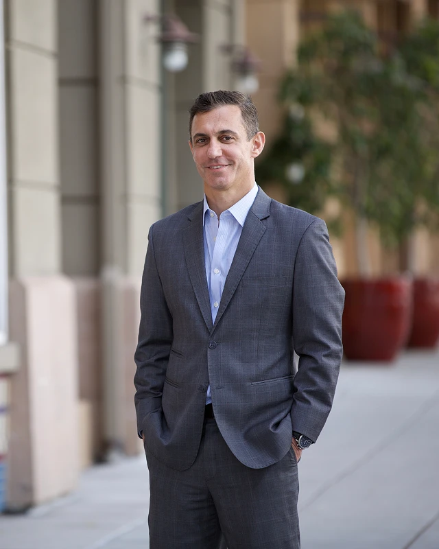 Man in a suit stands on a sidewalk with hands in pockets, smiling slightly