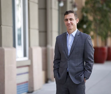 A man in a suit stands on a sidewalk near buildings and large planters