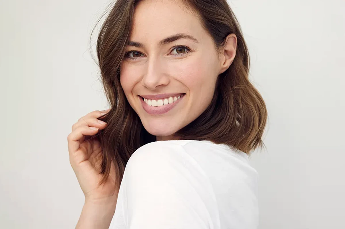 Woman with shoulder-length brown hair smiling and touching her hair. White background