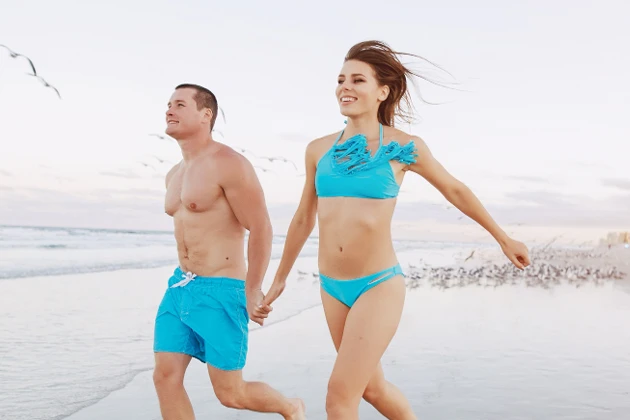 A man and a woman hold hands while walking on the beach wearing swimwear