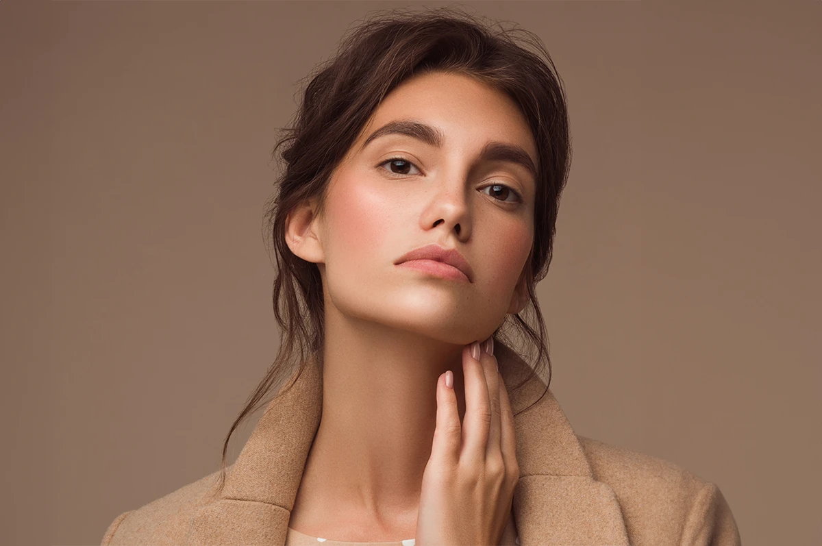 Close-up of a woman with long dark hair, wearing a beige coat, touching her neck