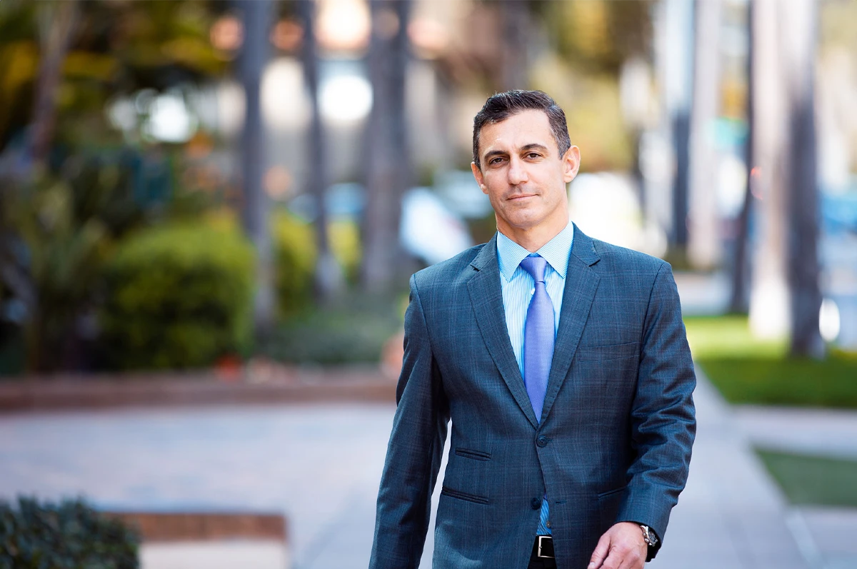 A man in a suit walks down a city sidewalk with greenery in the background