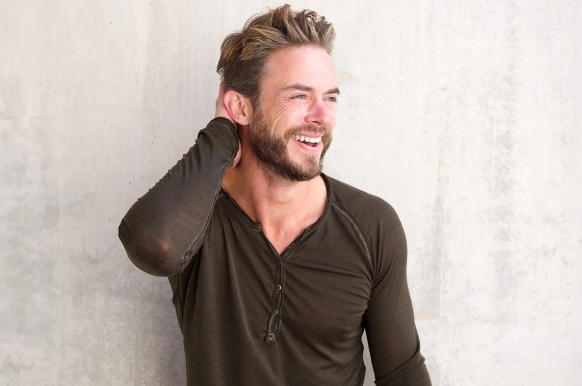 Smiling man with short, styled hair, wearing a long-sleeve shirt, standing against a wall