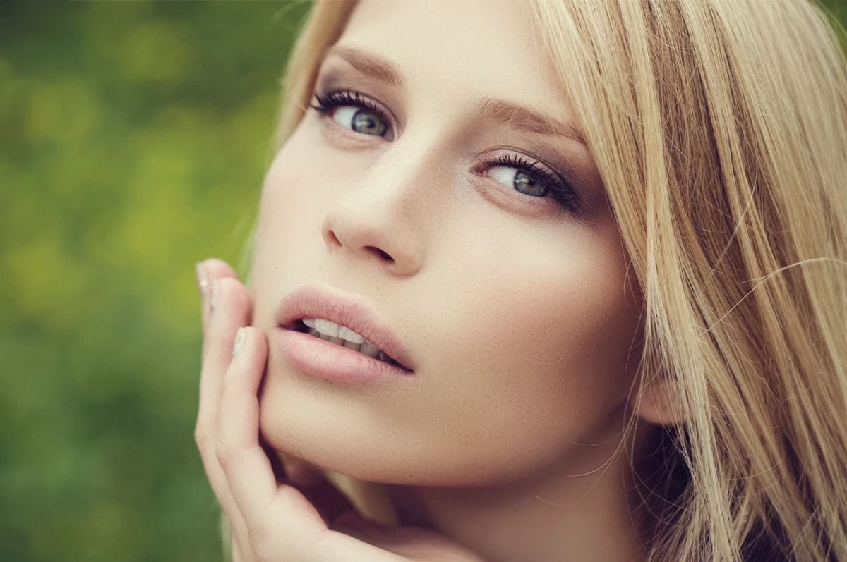 Close-up of a person with blonde hair, touching their face, against a blurred green background