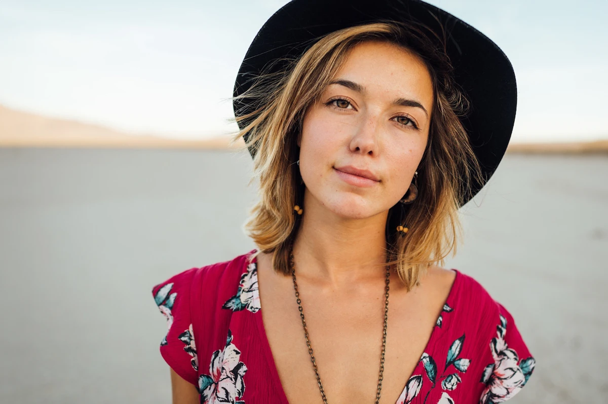 A woman with shoulder-length hair wears a black hat and a floral dress. She stands in a dry landscap