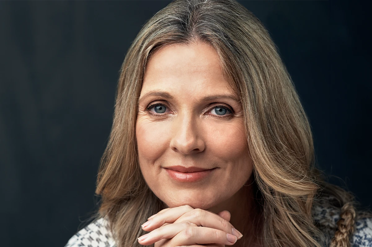 A woman with long, wavy hair smiles while resting her chin on her hands