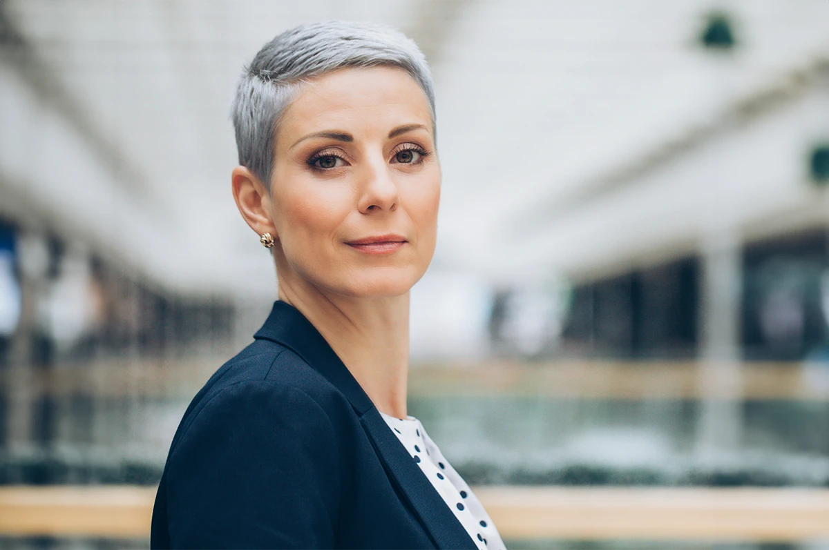 A woman with short gray hair, wearing a blazer, looks confidently at the camera