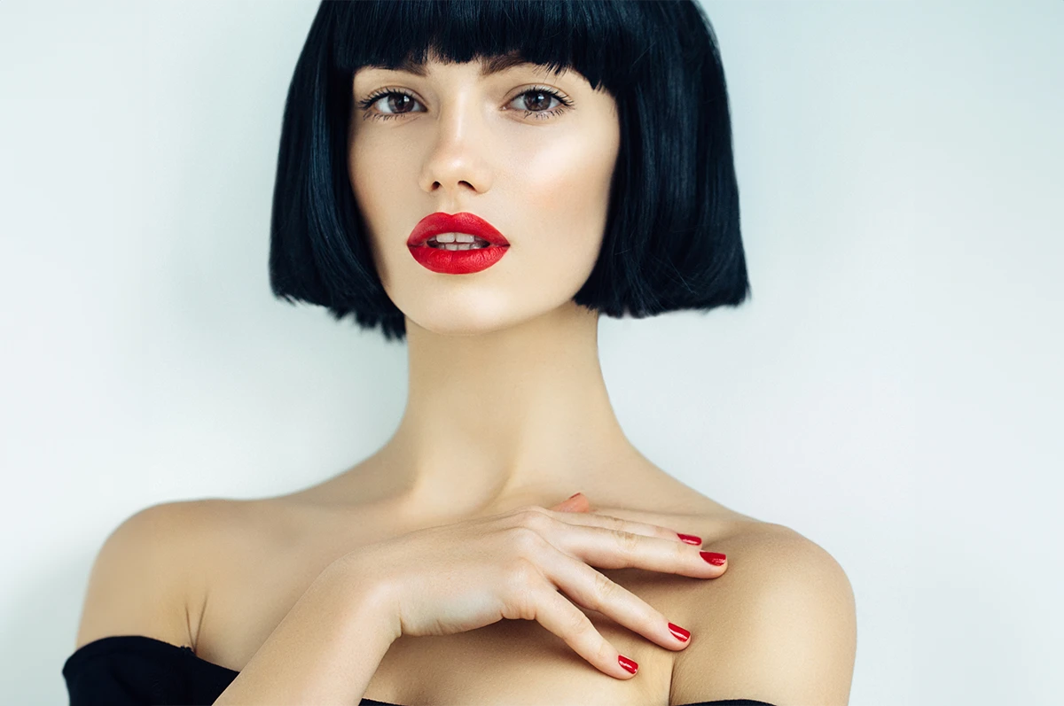 A woman with a bob haircut and red lips poses, wearing a black off-the-shoulder top