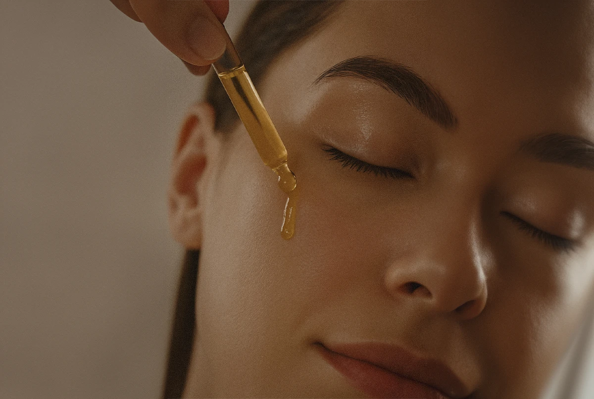 A person applying facial oil with a dropper on their cheek. Their eyes are closed