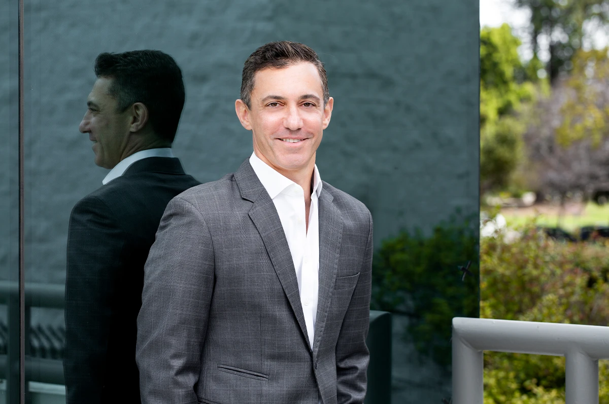 Man in a gray suit standing next to a reflective surface with greenery in the background