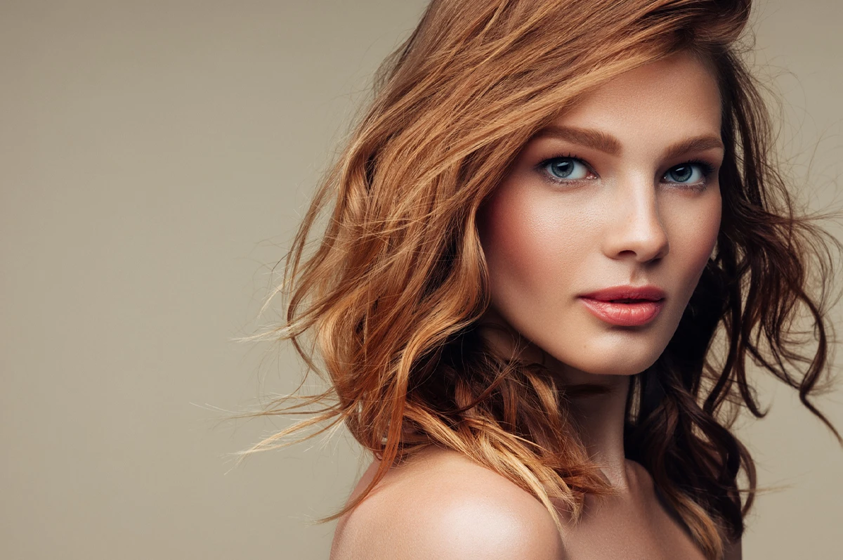 A young woman with wavy reddish-brown hair poses against a neutral background