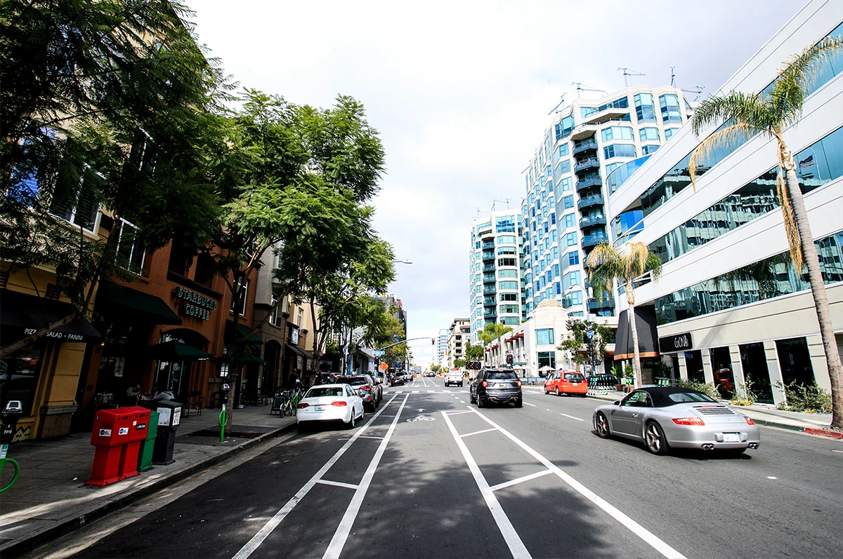 Street view with cars, trees, and buildings on either side under a cloudy sky