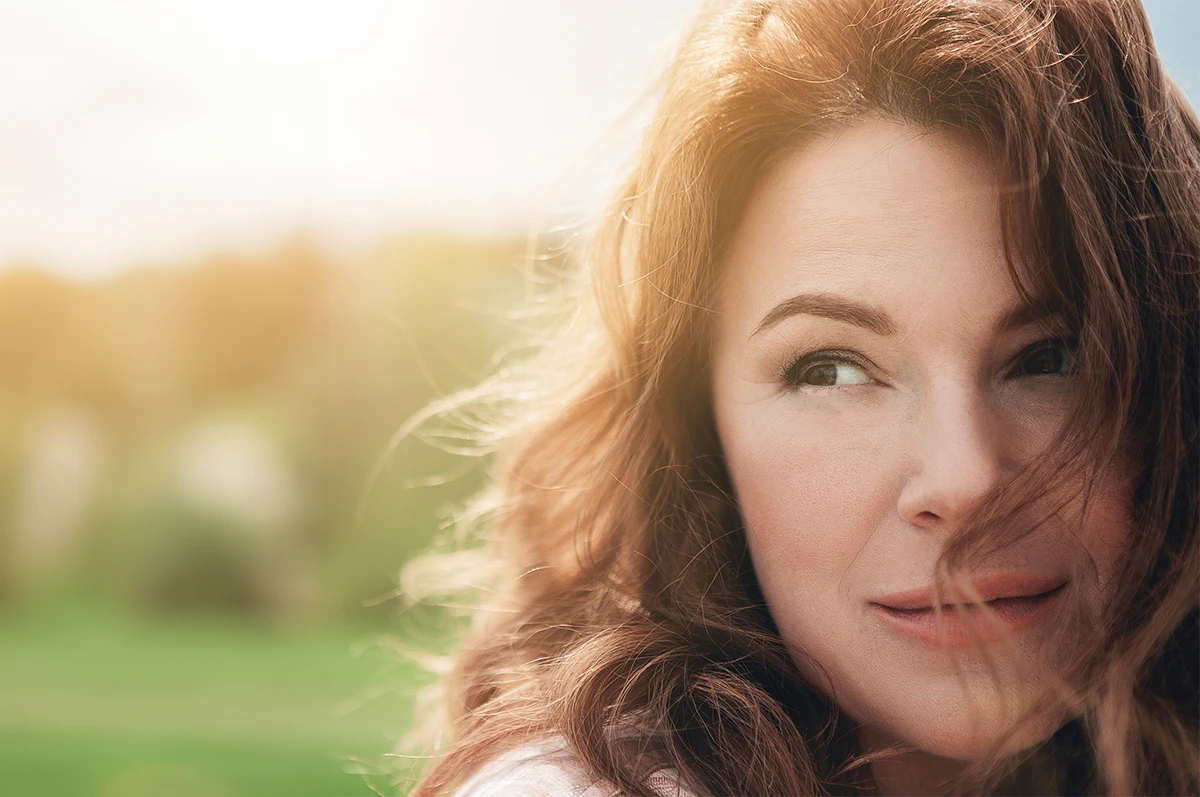 A woman with wavy brown hair looks away, with a blurred green background
