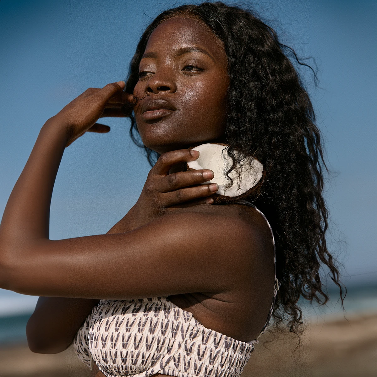 A woman with curly hair poses outdoors, holding a shell near her neck